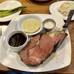 Prime Rib - A meal featuring a large prime rib steak with mashed potatoes, gravy, and dipping sauces, served on a white plate with side dishes and salad in the background.