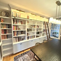 ACS Woodshop - Custom white built-in bookshelves with upper lighting, a sliding library ladder, and lower storage cabinets in a bright home.