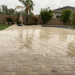 Travertine tile, San Miguel. Raining! - A wet outdoor patio features travertine tile flooring, with visible reflections from recent rain. The backyard includes landscaped plants, trees, and shrubs, with a pool visible in the background. There are decorative garden elements, such as benches and an umbrella stand, and a house with a tiled roof is seen beyond the yard. The scene captures a rainy day in a residential outdoor space.