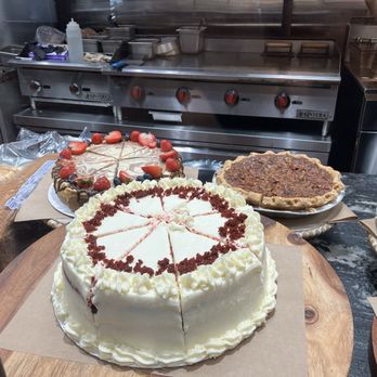 OAK'D BBQ - Cakes! - A variety of cakes are placed on a counter in a commercial kitchen or bakery environment. The prominent cake in the foreground is a white frosted cake with sliced pieces, decorated with white frosting and red crumbs, and topped with white frosting rosettes. Behind it, there are additional cakes, including one with strawberries and chocolate drizzle, and others with different toppings and crusts. The background features kitchen equipment, such as a stove and utensils, indicating a professional food preparation setting. - Barbeque Near Me - Addison, Texas