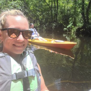 Kayaking St Augustine - A woman wearing sunglasses and a life jacket is taking a selfie on a river, with a yellow kayak and another person in the background. The river is surrounded by dense green trees, indicating a natural outdoor setting suitable for kayaking activities. - Rafting/Kayaking Near Me