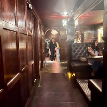 Constitution Bar - The image shows a dimly lit bar interior with dark wood paneling on the walls and ceiling. There are booths with tufted leather seats along the right side, and a corridor leading to the bar area in the background. Patrons and staff are visible, with a woman sitting in a booth on the right and a staff member standing near the bar. Chandeliers and decorative lighting add to the ambiance. - Speakeasies Near Me - Lexington, Kentucky
