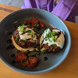 Crab Cakes - Fried breaded dishes topped with sauce and herbs are presented with cherry tomatoes on a gray plate, likely at a restaurant.