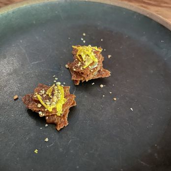 Uchi Austin - The amouse bouche at the beginning (little cracker thing, it's got some good flavor) - Two small food pieces with yellow zest garnish are placed on a black plate, with a wooden table in the background. - Sushi Bars Near Me - Austin, Texas