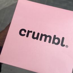 A pink Crumbl cookie box is held by a person, with a gray tiled floor in the background.