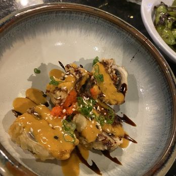 Tsukimi - Stuffed Mushroom - A sushi roll is presented on a ceramic plate, topped with multiple sauces, chopped green onions, and orange fish roe. The roll appears to be tempura-fried or battered, with additional drizzles of dark and light sauces. In the background, there are side dishes, including a bowl of salad with leafy greens. The setting suggests a Japanese culinary establishment offering sushi and related dishes. - Sushi Bars Near Me - Austin, Texas
