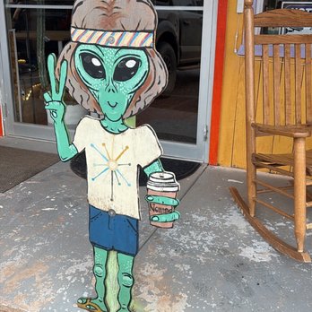 Nomad Cafe - A colorful wooden cutout of an alien with large black eyes, wearing a headband, t-shirt, shorts, and flip-flops, holding a coffee cup and making a peace sign outside a cafe. - Coffee & Tea Near Me - Kanab, Utah