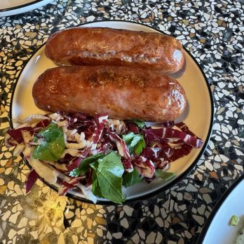 Fulano - Argentinean Chorizo - Two grilled sausages are placed on a plate alongside a mixed salad with leafy greens and radicchio, on a speckled tabletop. - Argentine Near Me - Key Biscayne, Florida
