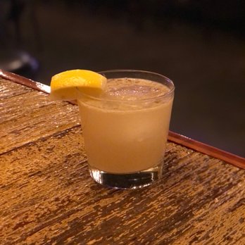 Free State - The good doc ($10) - A clear glass containing a light-colored cocktail with ice is positioned on a textured wooden bar surface. A lemon wedge is used as a garnish on the rim of the glass. The background is dark and out of focus, indicating a nighttime or low-light environment, likely at a bar or restaurant. - Wine Bars Near Me - Washington, DC, District of Columbia