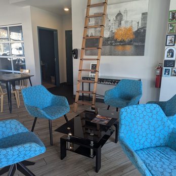 Epic Coffee - The photo depicts the interior of a coffee shop featuring four blue patterned chairs arranged around a black coffee table. A large black-and-white cityscape artwork with a tree is mounted on the wall behind the chairs. A wooden ladder is positioned against the wall, leading to a small window or storage space above. The floor has wood-like tiles, and part of a dining area with additional tables and chairs is visible. The setting appears cozy and modern. - Juice Bars & Smoothies Near Me - Carmel, Indiana