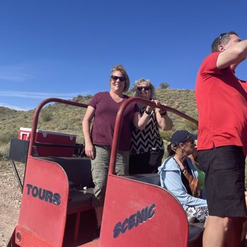 Apache Trail Tours - Four people are on a guided off-road tour vehicle in a desert landscape with hills and sparse vegetation. Two women are standing, one smiling and the other taking a photo, while a man in a red shirt is taking a selfie. The vehicle is red, with the words 'TOURS' and 'SCENIC' visible on its side. The scene is set under a clear blue sky, indicating a sunny day. - Tours Near Me - Apache Junction, Arizona