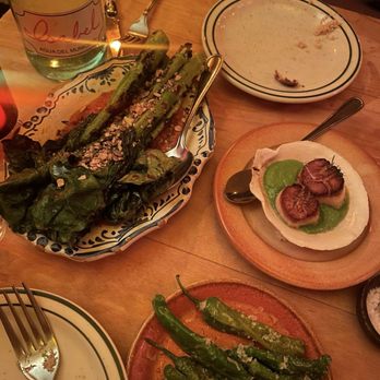 Bar Isabel - The image shows a wooden table with various dishes including grilled green peppers, a scallop dish with green sauce, and grilled asparagus, along with a bottle of water. - Spanish Near Me - Toronto, Ontario