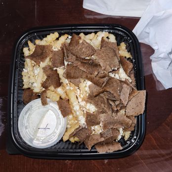 Gyro Shack - Greek fries with gyro meat. Meal in itself. - A black plastic takeout container features mashed potatoes topped with sliced roast beef. The container is placed on a dark wooden table, with a black fork, a white napkin, and a crumpled paper napkin nearby. The mashed potatoes appear creamy, and the roast beef slices are thin and cooked. A small clear plastic cup with gravy is also inside the container. - Greek Near Me - Albuquerque, New Mexico