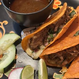 3 Piece Birria Tacos - Three birria tacos filled with shredded beef, garnished with onions and cilantro, served with lime, cucumber, radish, and a cup of broth.