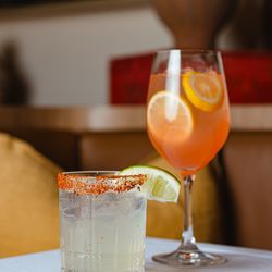 Felice Brickell - Two refreshing cocktails: a rocks glass with a Tajin rim and lime garnish next to a tall wine glass filled with an orange-hue...