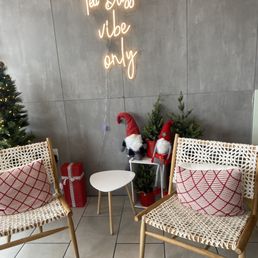 The scene features two woven chairs with red patterned pillows positioned on a tiled floor. Between the chairs, there are small Christmas trees and gnome decorations with red hats. A neon sign on the gray wall above reads 'Ta Bliss vibe only.' The setting appears to be a decorated indoor space, possibly a waiting or lounge area with holiday decor.