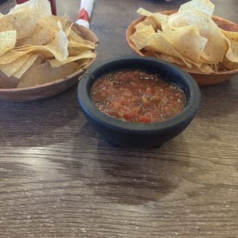 Los Gringos Locos - Chips and salsa - Two bowls of tortilla chips are positioned on a wooden table, with a small black dish of salsa in the center. One bowl contains plain tortilla chips, while the other has slightly thicker chips. A person's tattooed arm is visible in the background, along with a red drink and a napkin with a utensil. The setting appears to be a casual dining restaurant. - Mexican Near Me - Apache Junction, Arizona