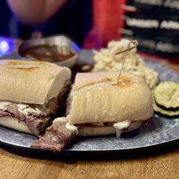 French dip - A French dip sandwich featuring sliced roast beef and melted cheese on a toasted bun is presented on a metal tray. The tray also includes sliced pickles, potato salad, and a small container of dipping au jus sauce. The background shows a dimly lit restaurant setting with a chalkboard menu partially visible.