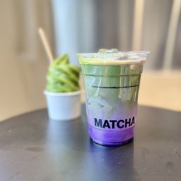 Blu and soft serve - A colorful matcha drink in a transparent cup features a swirling pattern of purple and green, with the label 'MATCHA' on the front. The drink is placed on a dark, smooth table, with a blurred background that includes a small cup with a green soft serve or whipped topping and a wooden stir stick. The setting appears to be indoors, possibly in a cafe or restaurant.