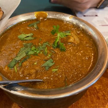 Spice House - Goat curry - A metal bowl contains goat curry with a rich, reddish-brown sauce, garnished with cilantro leaves, on a restaurant table. - Indian Near Me - Edison, New Jersey