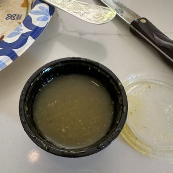 Los Taquitos - Salsa Verde - A black plastic container filled with green salsa sits on a white table, with a clear plastic lid nearby. A knife and a decorative spoon are also visible. - Mexican Near Me - Phoenix, Arizona
