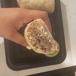 A person holds a partially eaten burrito filled with rice, meat, and vegetables, with a whole burrito on a black tray in the background.