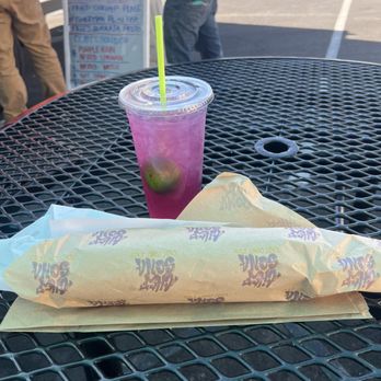 Chef Sena East Coast Eats - Chef Sena  has the best Philly cheesesteak I've ever tasted in a very long time. He is in Grubhub downtown Modesto. - A pink-colored drink with lime slices inside and a green straw is placed on a black metal outdoor table. Next to it is a wrapped sandwich in branded paper, likely from a fast-food or casual dining establishment. The background shows a parking lot with a menu board and two people standing nearby, with sunlight casting shadows on the scene. - Caterers Near Me - Modesto, California