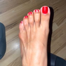 A close-up of a foot with red nail polish on the toenails, positioned on a wooden floor, with part of a shoe visible.