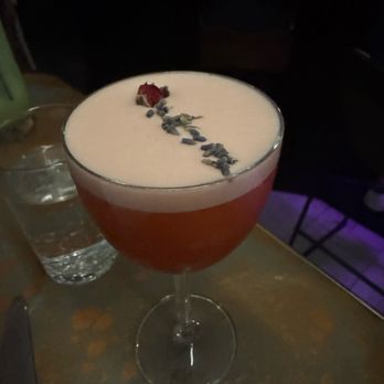 Ramro - A cocktail in a coupe glass with a frothy top, garnished with a dried flower and herbs, on a table with a glass of water nearby. - Himalayan/Nepalese Near Me - Astoria, New York