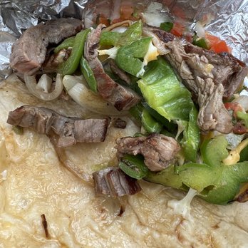 Mr Chingon - Beef Fajita Taco - The image shows a beef fajita taco with grilled beef strips, sautéed green peppers, onions, and melted cheese inside a soft flour tortilla. The taco is partially wrapped in aluminum foil, with some of the toppings spilling out. A small container of sauce or dressing is visible in the background, placed on a wooden surface. - Tacos Near Me - Austin, Texas