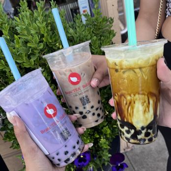 BeauTea Bubbles - Taro milk tea, matcha brown sugar and lavender milk tea with boba - Three bubble tea drinks with tapioca pearls are held by different individuals outside a tea lounge. The drinks include a purple milk tea, a beige milk tea with black tapioca pearls, and a yellow and brown swirled tea with ice. Each cup has a colored straw, and the background features green plants and signage indicating payment options and the business location. The setting appears to be outside a tea lounge or bubble tea shop. - Bubble Tea Near Me - Denver, Colorado