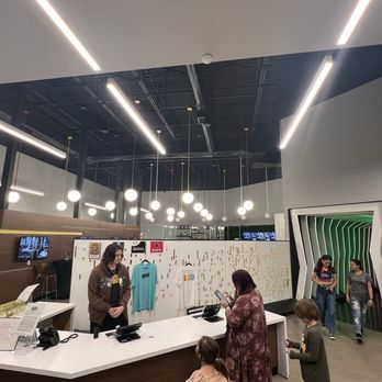 Electric Playhouse - A spacious, contemporary entertainment venue features a reception desk, hanging lights, and a green-lit entrance. Several people are present, some using phones, others waiting or walking. - Party & Event Planning Near Me - Albuquerque, New Mexico