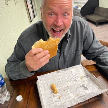Bodega On Smith - Cubano and a happy hubs - A man with gray hair and a beard is sitting at a wooden table, holding a sandwich and smiling widely. There is a newspaper on the table and a water bottle nearby. - American Near Me - Providence, Rhode Island