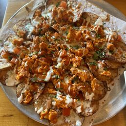 Buffalo chicken nachos - A serving of buffalo chicken nachos features potato chips topped with shredded chicken, melted cheese, diced tomatoes, and garnished with herbs. The dish is drizzled with white and brown sauces, presented on a metal tray lined with parchment paper, with a wooden table surface visible in the background.