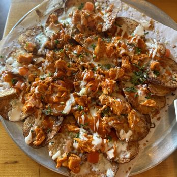 Commonwealth - Buffalo chicken nachos - A serving of buffalo chicken nachos features potato chips topped with shredded chicken, melted cheese, diced tomatoes, and garnished with herbs. The dish is drizzled with white and brown sauces, presented on a metal tray lined with parchment paper, with a wooden table surface visible in the background. - Beer Bar Near Me - Modesto, California