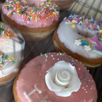 King Donuts - Multiple donuts with various icing colors and decorations are shown on a wire rack. One donut has pink icing with multicolored sprinkles and the word 'I ♥ U' written on it. Another donut features white icing with a rainbow and clouds, while others have pink or white icing with sprinkles. The donuts are arranged closely together, highlighting their decorative toppings and festive appearance. - Donuts Near Me - Temecula, California