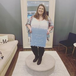 A woman with shoulder-length hair is standing on a round ottoman, holding a sign that reads 'I got my something new at Something Blue.' She is smiling and positioned in front of a large mirror in a room with a beige sofa and a black chair.