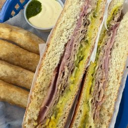 Cubano - A Cuban sandwich with layers of ham, pork, cheese, and pickles is served on a bun, accompanied by fried yuca sticks and a small cup of green sauce.