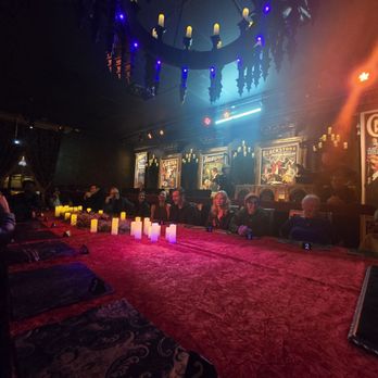 Mystique The Magic Experience - A group of people sit around a long table in a dark, atmospheric room decorated with vintage posters and candle-like lights. - Magicians Near Me - Mesa, Arizona