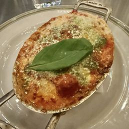 Baked Eggplant Parm - A baked eggplant parmesan dish features a breaded and baked eggplant slice topped with melted cheese and tomato sauce, garnished with a fresh basil leaf. The dish is presented on a metal tray with handles, placed on a white plate. In the background, there are glasses of water on a wooden table, indicating a restaurant setting.