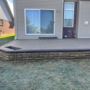 Unique Cement - The image shows a concrete patio with a textured surface, built adjacent to a residential house. The patio features a stone veneer border along its edge, with a small built-in planter or feature on the left side. The house has beige siding, a sliding glass door, and multiple windows. The yard surrounding the patio is grassy, with neighboring houses visible in the background. The scene appears to be part of a home improvement or landscaping project. - Masonry/Concrete Near Me - Clinton Township, Michigan