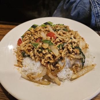 Thai Kitchen - Thai Basil Chicken - A white plate contains Thai basil chicken with ground chicken, onions, cherry tomatoes, and basil leaves, served over a bed of white rice. The dish is presented on a wooden table, with a person wearing denim jeans visible in the background. A smartphone and a napkin are also on the table. - Thai Near Me - Austin, Texas