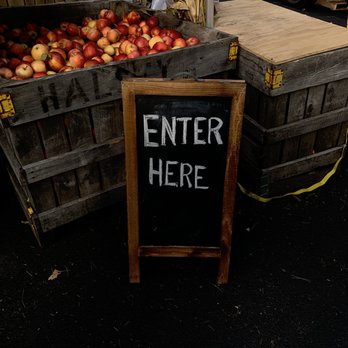 Jericho Cider Mill - A chalkboard sign with a wooden frame displays the words 'ENTER HERE' in white chalk. It is placed on a dark, textured ground in front of large wooden crates filled with red and yellow apples. The market setting includes additional wooden platforms and a truck visible in the background, indicating an outdoor produce stand or farmers market. - Fruits & Veggies Near Me - Jericho, New York