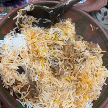 Clay Handi - Biryani - A large, round clay bowl contains biryani rice mixed with pieces of meat, vegetables, and spices. A wooden spoon rests inside the bowl, used for serving. The dish appears to be part of a traditional meal, with visible rice grains, meat chunks, and some vegetable pieces, reflecting authentic Pakistani or Indian cuisine. - Halal Near Me - Buffalo, New York