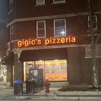 Gigio's Pizzeria - Stop in if you see this - The image shows the exterior of Gigio's Pizzeria, a brick building with a large illuminated orange sign displaying the business name. The storefront has a window revealing the interior menu and seating area. Outside, there is a trash can, a fire hydrant, and a small landscaped area with a tree. The scene appears to be taken at night, with streetlights and interior lighting illuminating the area. - Pizza Near Me - Evanston, Illinois