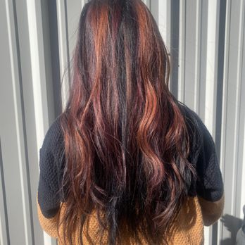 Studio 80 Salon - A woman with long, wavy hair with multiple shades of red, black, and brown stands outdoors in front of a metal wall, with sunlight illuminating her hair. - Waxing Near Me - Patchogue, New York
