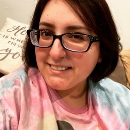 A woman with short dark hair, glasses, and a nose ring is smiling at the camera. She is wearing a tie-dye shirt and is indoors, with a pillow in the background.