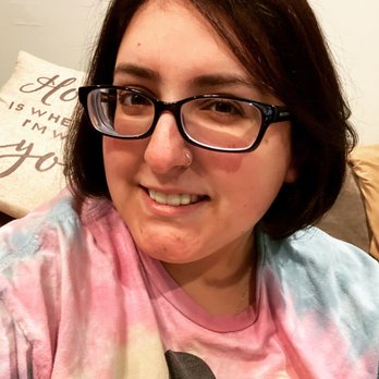 Studio 80 Salon - A woman with short dark hair, glasses, and a nose ring is smiling at the camera. She is wearing a tie-dye shirt and is indoors, with a pillow in the background. - Waxing Near Me - Patchogue, New York