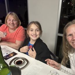 Girls Pre-birthday dinner with nana/mama in town! - Three women and a girl are seated at a table in a restaurant, smiling for a selfie. The table has a bottle, soy sauce, and menus, with a dark indoor background.