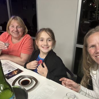 Sushi Bene - Girls Pre-birthday dinner with nana/mama in town! - Three women and a girl are seated at a table in a restaurant, smiling for a selfie. The table has a bottle, soy sauce, and menus, with a dark indoor background. - Japanese Near Me - Prosper, Texas